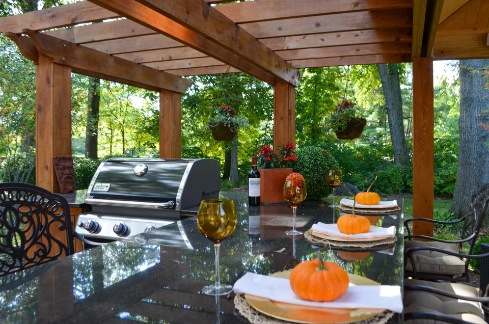 Fall dining table under pergola framing with grill, hanging planters, wood bar, paver patio, and glassware.